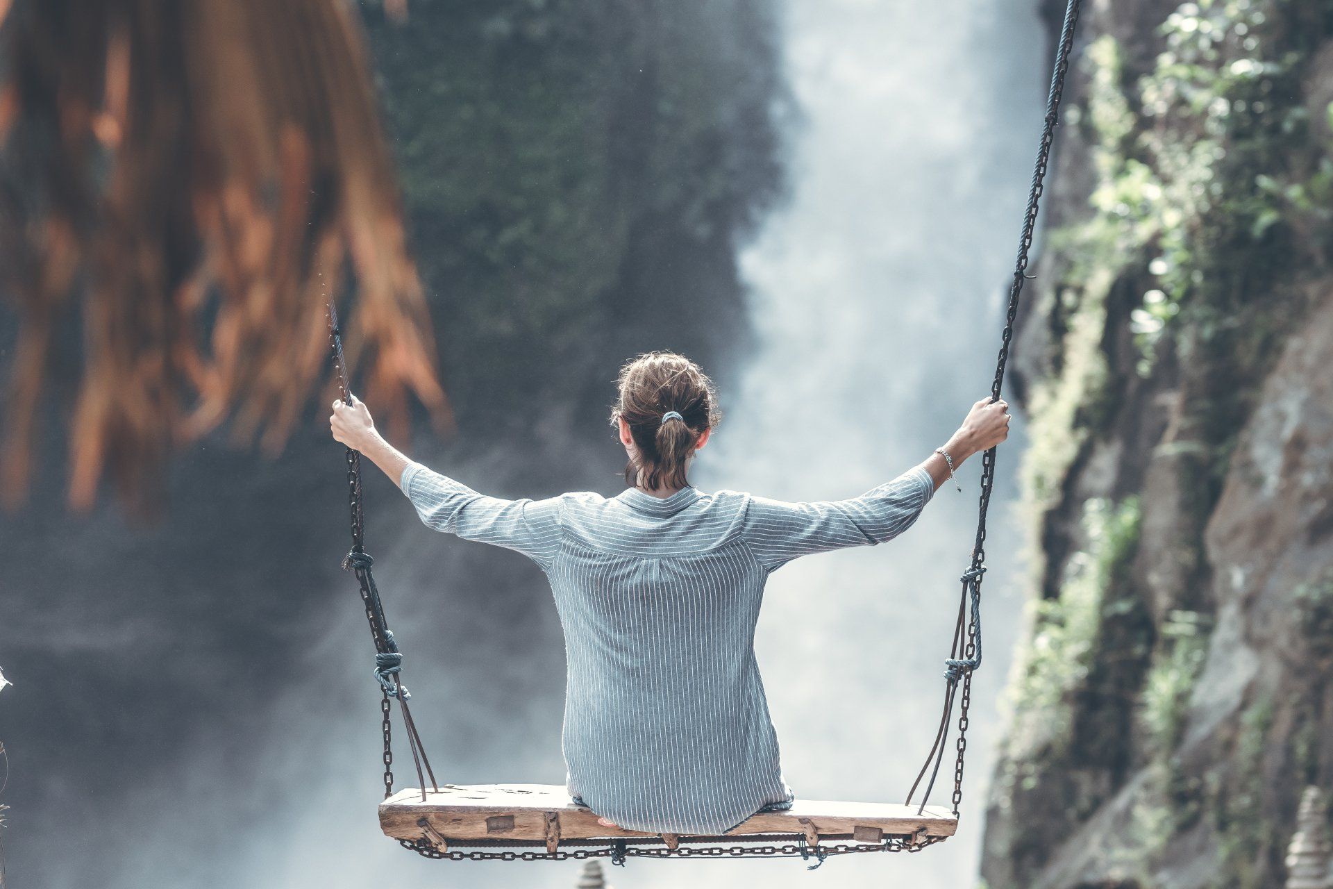 Person sitting on a rope swing facing a misty waterfall canyon