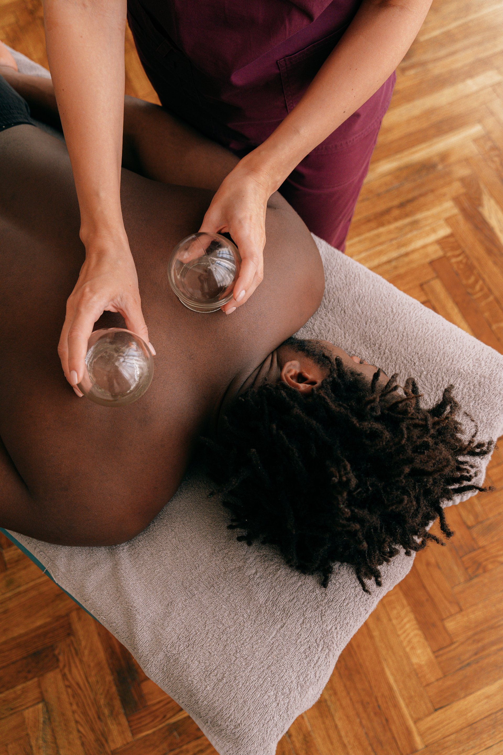 Person receiving back massage with hot stones on a gray towel beside a wooden floor